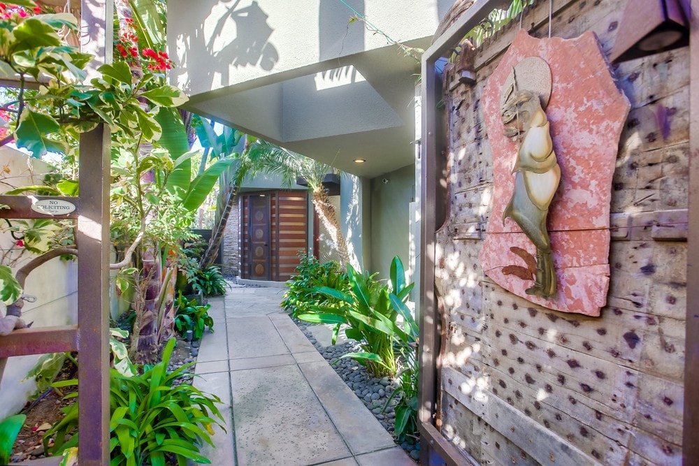 A tranquil pathway leads to the entrance, flanked by lush greenery and vibrant flowers. A uniquely crafted hanging ornament adorns the wall, adding artistic charm. Soft light filters through the foliage, creating a serene ambiance around the entryway.
