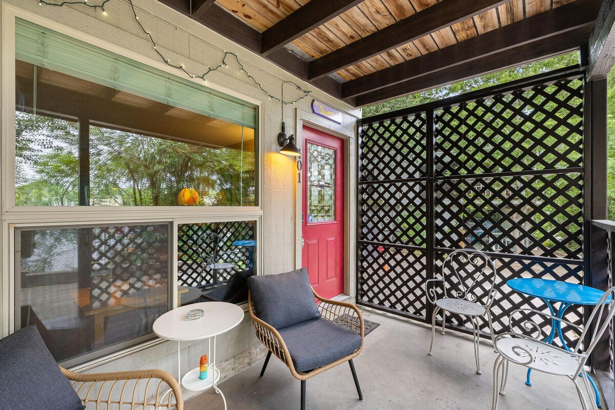 A covered patio area is presented, featuring a cozy seating arrangement with two dark chairs and a small white table. String lights hang along the ceiling. A bright red door with a decorative screen is positioned on one side, adding a welcoming touch.