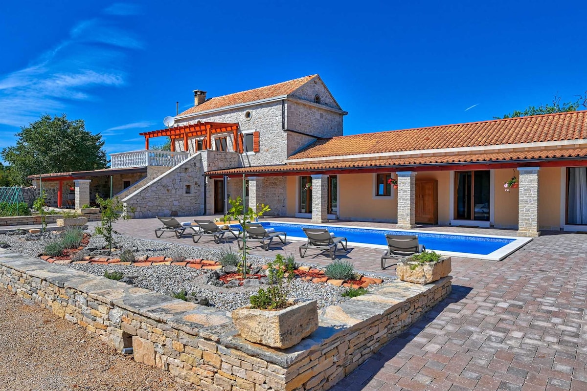 A spacious villa showcases a large swimming pool surrounded by a landscaped courtyard. Comfortable lounge chairs are positioned by the pool, and the building features a traditional stone facade with a terracotta roof. Green plants and decorative stones enhance the outdoor setting.