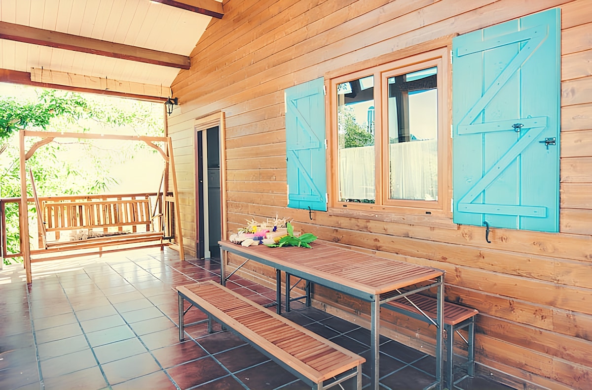 An outdoor space is showcased with a wooden table and benches, complemented by a swing hanging nearby. Bright blue shutters frame two windows, allowing natural light to enter. The tiled floor adds a welcoming touch to the area, perfect for meals or relaxation.