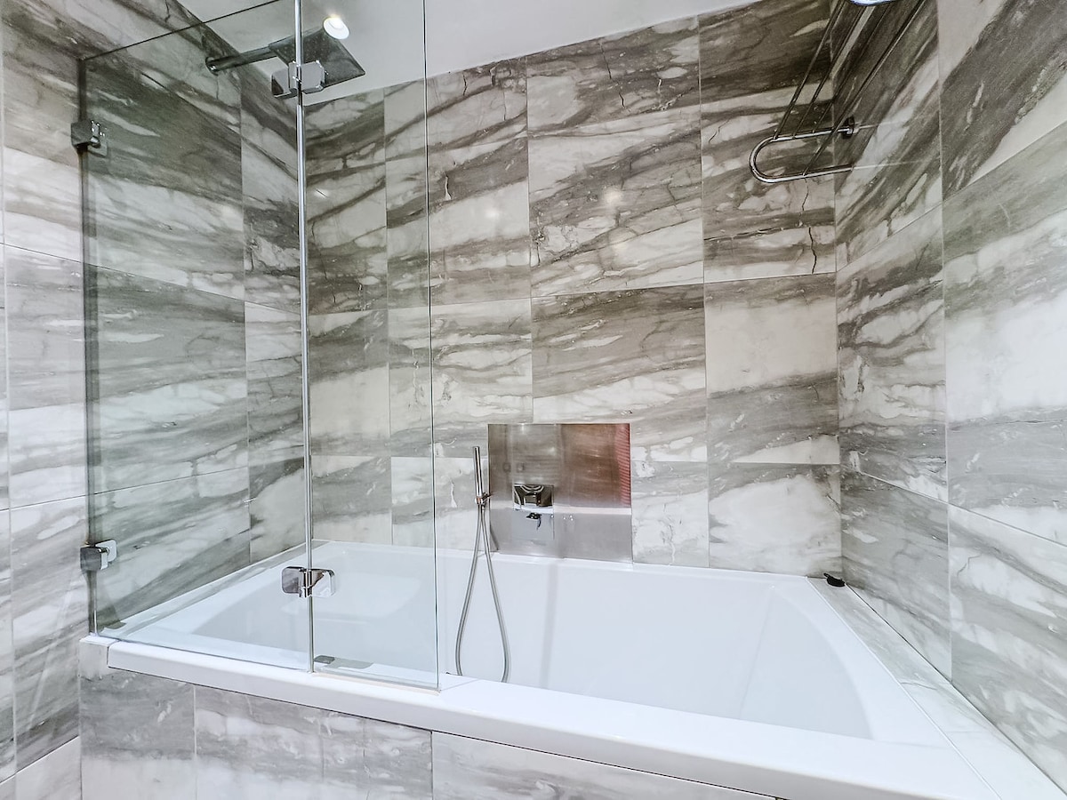 A modern bathroom features a glass-enclosed bathtub with a sleek design. Subtle gray and white marble accents adorn the walls, enhancing the sophisticated appearance. Fixtures and a handheld shower are positioned within easy reach, providing convenience for relaxation.