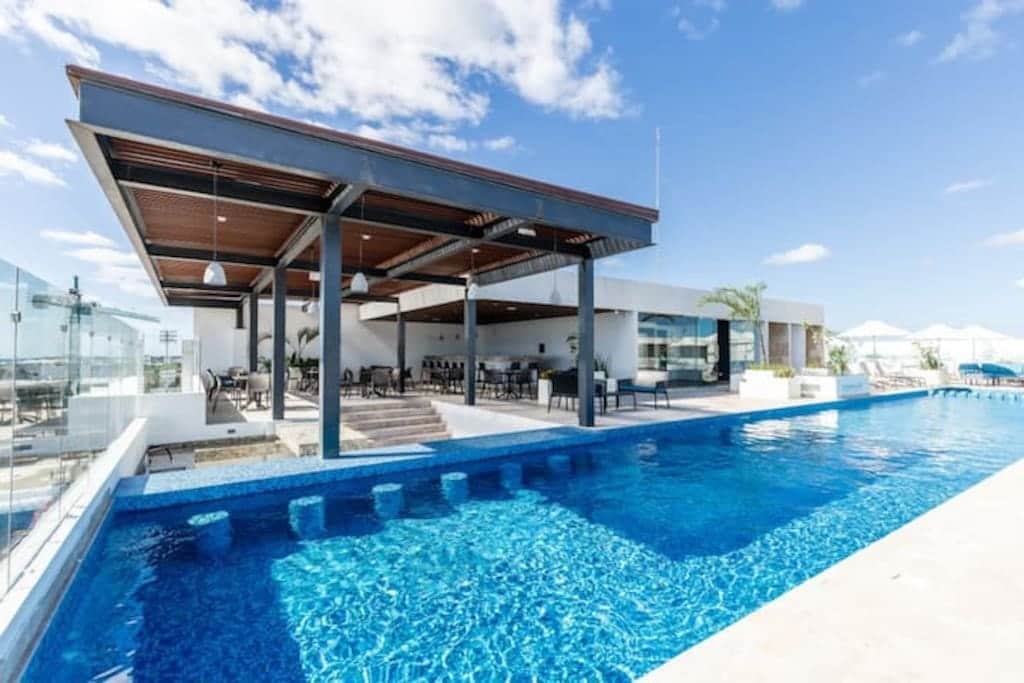 An expansive rooftop pool area is seen, featuring a shimmering blue pool under a clear sky. Comfortable seating arrangements are provided under a stylish shaded structure, surrounded by lush greenery and sun loungers, offering a tranquil space for relaxation and enjoyment.