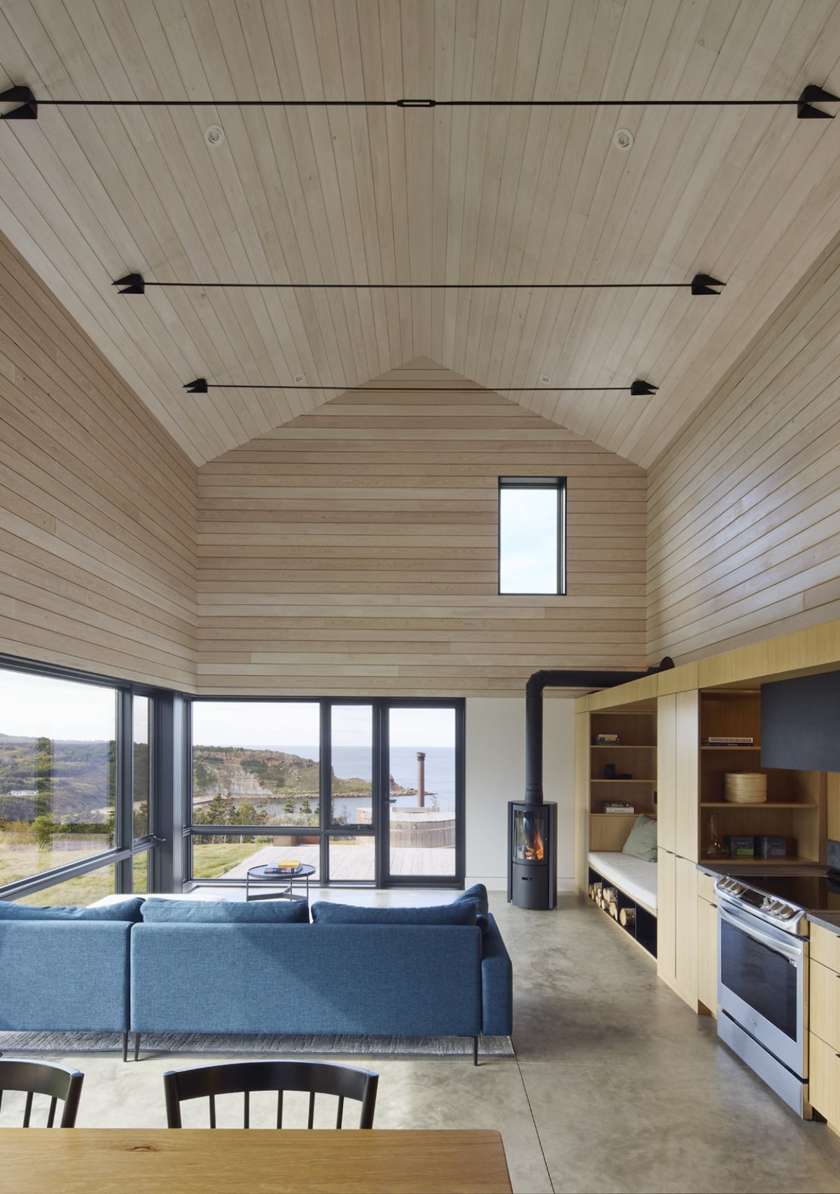 The interior of Birch Cottage showcases a spacious living area with high wooden ceilings. Large windows provide views of the outdoors, while a comfortable blue sofa is positioned near a modern wood stove. A well-equipped kitchen is visible, featuring light wood cabinetry.