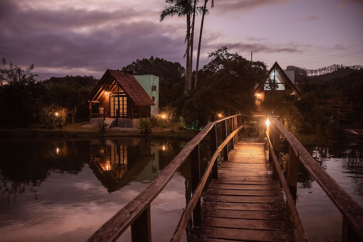 A wooden bridge leads to two charming cabins illuminated softly by outdoor lights. The tranquil water reflects the evening sky and surrounding trees, creating a serene environment. The reflections of the cabins enhance the peaceful ambiance of the natural setting.