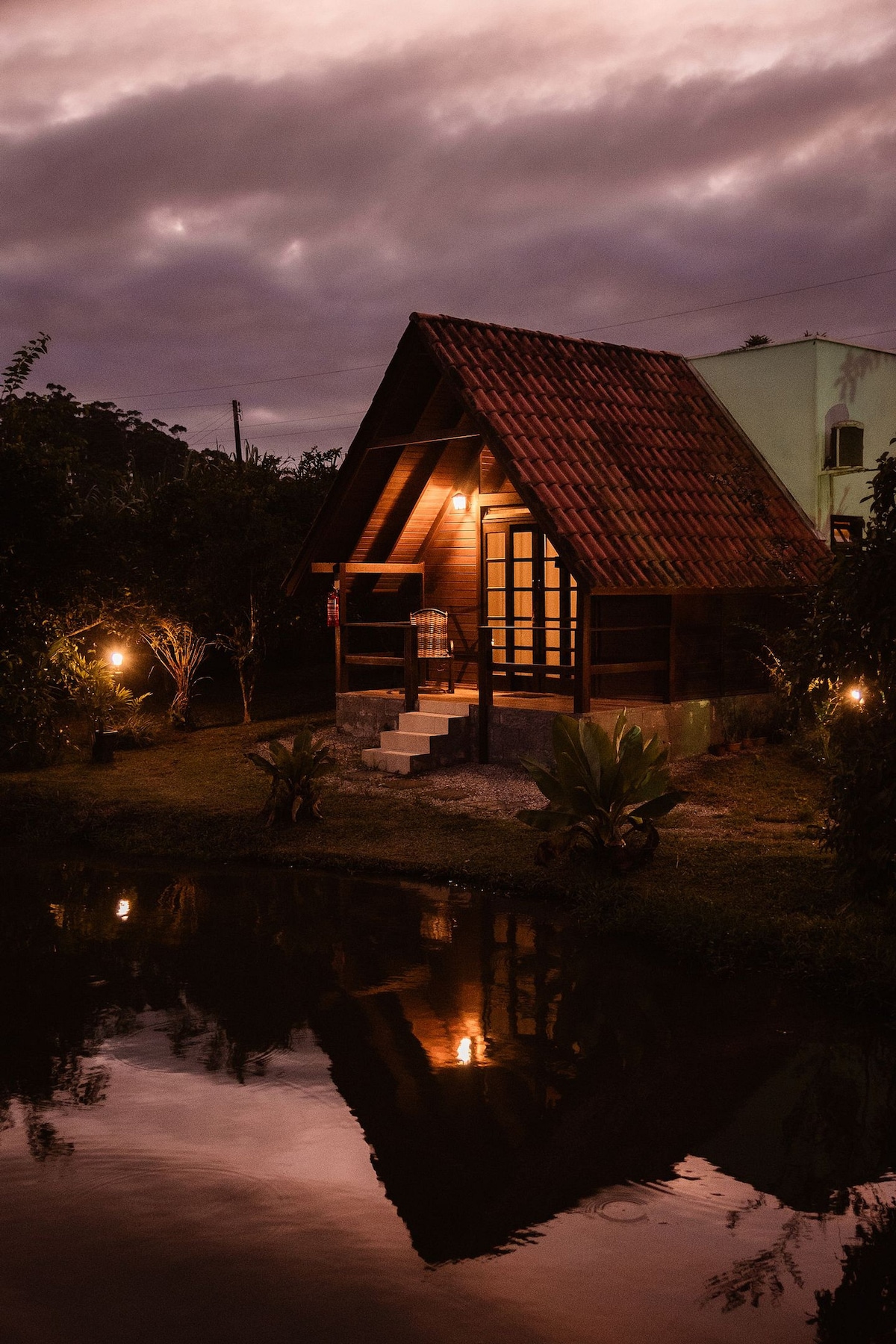 A charming chalet stands beside a tranquil pond, reflecting its wooden structure and sloped roof. Soft lighting highlights the entrance, surrounded by lush greenery and trees. A peaceful atmosphere is created by the scenic view of the darkening sky and the surrounding natural beauty.