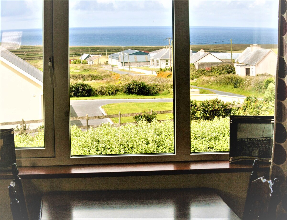 A large window frames a view of the Atlantic Ocean and surrounding coastal landscape. The scene includes colorful buildings and a winding road, with greenery in the foreground, allowing for a bright and airy atmosphere.