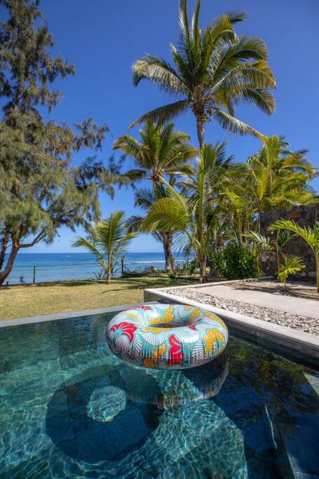 A colorful inflatable ring floats in a sparkling swimming pool, surrounded by lush tropical foliage and palm trees. The shimmering blue water reflects the clear sky and the distant view of the Indian Ocean enhances the tranquil setting.