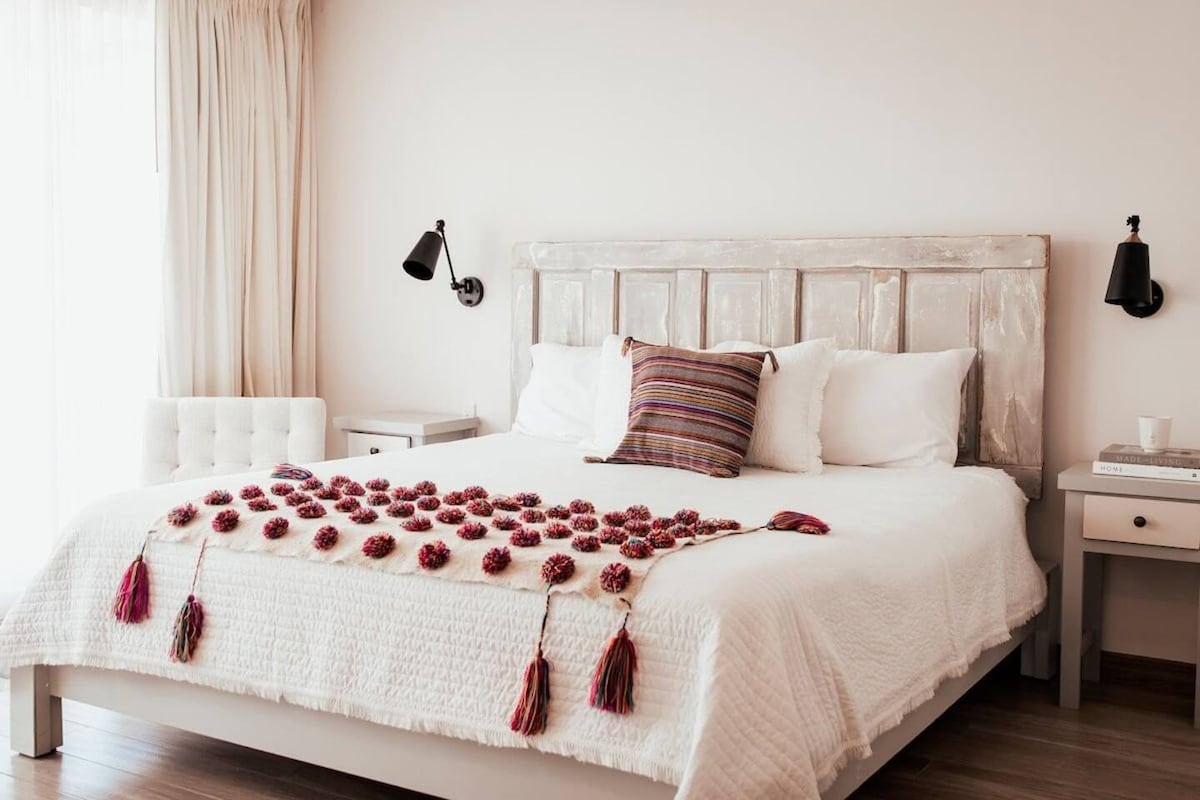A bright bedroom features a spacious bed with a textured white coverlet; a decorative arrangement of red tassels and a patterned pillow add visual interest. Light is softly filtered through sheer curtains, while wall-mounted lamps provide additional illumination on either side of the bed.
