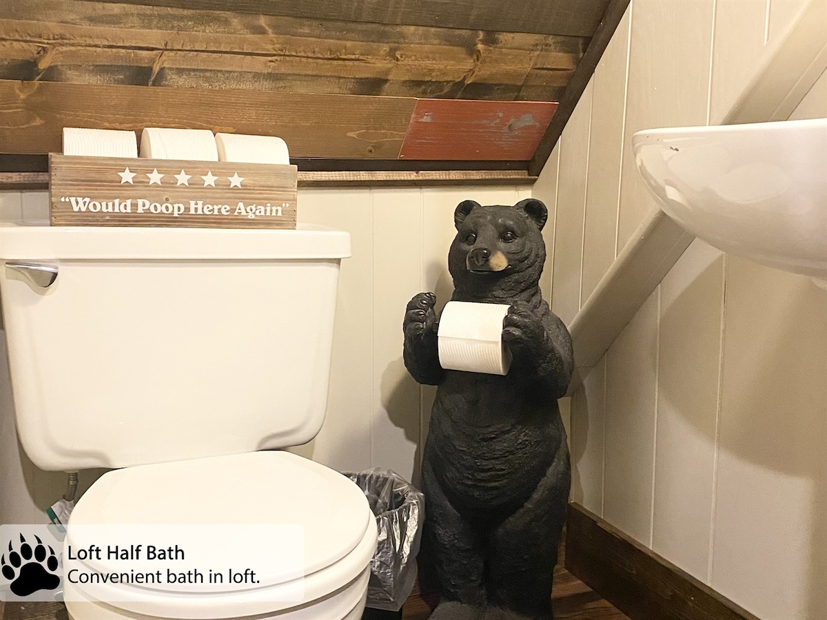 The half bath in the loft features a compact toilet alongside a bear statue holding extra toilet paper. A modern sink is positioned nearby, with wood accents on the walls adding a rustic touch. Essential amenities are provided for convenience.