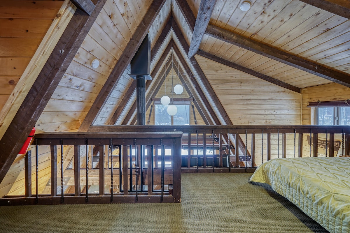 An A-frame structure features vaulted wood ceilings, showcasing exposed beams and large triangular windows. A cozy sleeping area is visible with a quilted cover on the bed. Soft lighting from pendant fixtures provides illumination, while a balcony railing adds to the open feeling of the space.