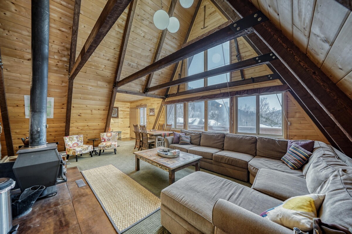 A spacious living area features high vaulted ceilings with exposed wooden beams. Large windows allow natural light to fill the space, highlighting a comfortable sectional sofa and a rustic coffee table. A wood-burning stove adds warmth, complemented by a cozy area rug.