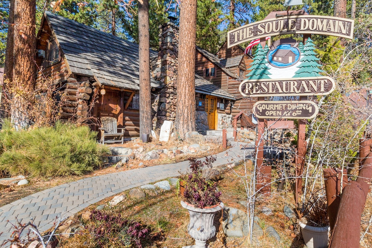 The exterior of a rustic cabin is framed by tall pine trees, with a stone pathway leading to the entrance. A sign identifies the establishment as 'The Soule Domain Restaurant,' featuring inviting wooden accents. Planters with minimal greenery enhance the welcoming atmosphere.