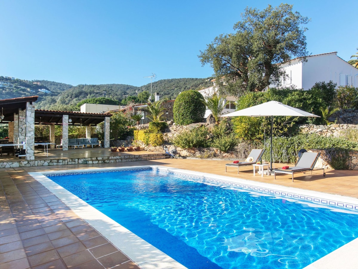 A sunlit swimming pool is framed by a spacious terrace featuring deck chairs and an umbrella for shade. Lush greenery and a stone structure are visible in the background, creating a serene outdoor space for relaxation.