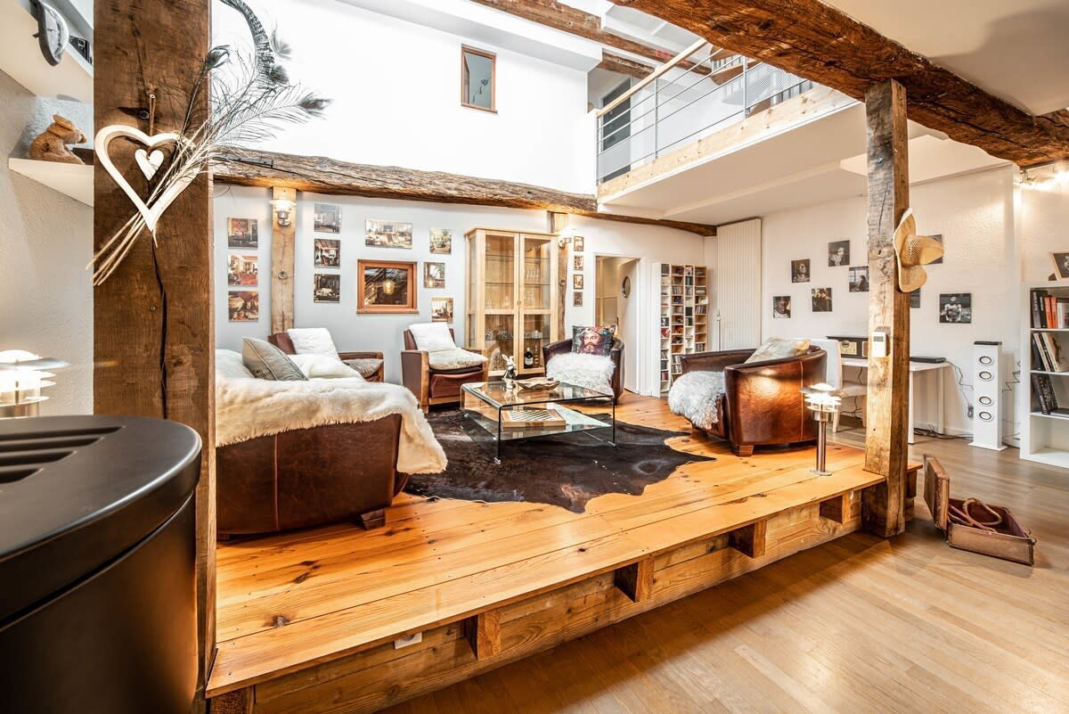 A spacious living area features a wooden floor with a sunken seating area. Two armchairs and a large sofa, draped with cozy throws, create a relaxed atmosphere. Large windows allow natural light to illuminate the room, complemented by wooden beams and decorative wall art.