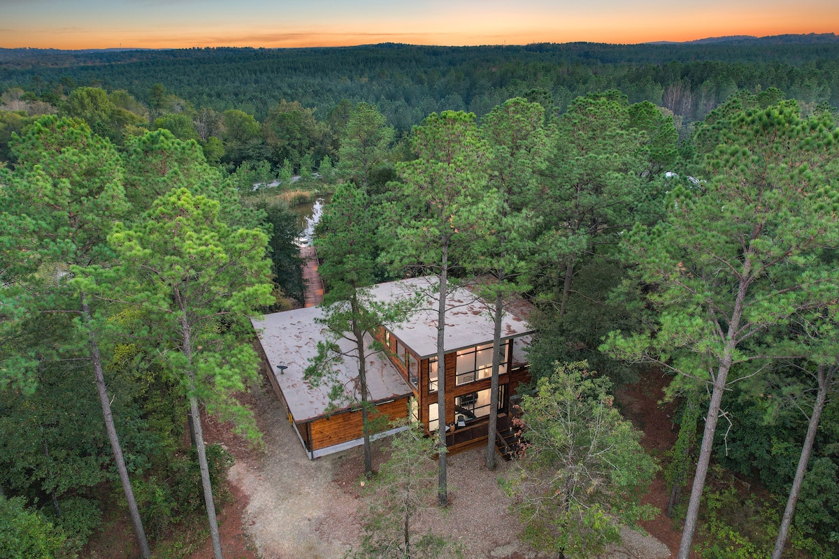 The cabin is nestled among tall pine trees, surrounded by lush greenery. Its modern design features large windows that reflect the colors of the sunset, while natural materials blend harmoniously with the landscape. A winding pathway leads through the trees to the cabin's entrance.