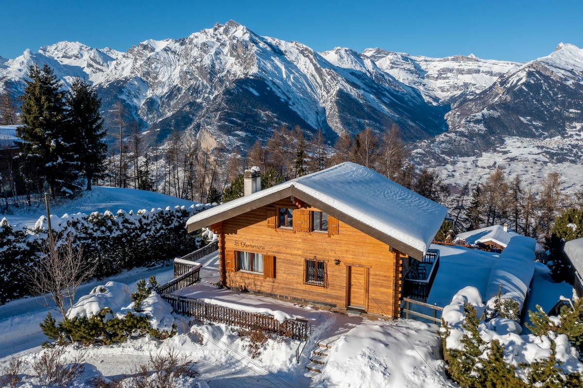 The wooden chalet is surrounded by snow-covered mountains, offering a panoramic view of the valley below. The exterior features a balcony and terraces that provide a serene outdoor space. Tall trees frame the property, enhancing the natural setting.