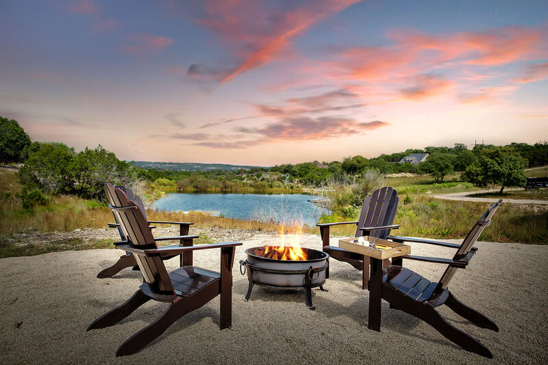 A circle of four wooden Adirondack chairs surrounds a fire pit, with a glowing fire at its center. The scene is complemented by a serene body of water in the background, flanked by lush greenery under a colorful sunset sky.