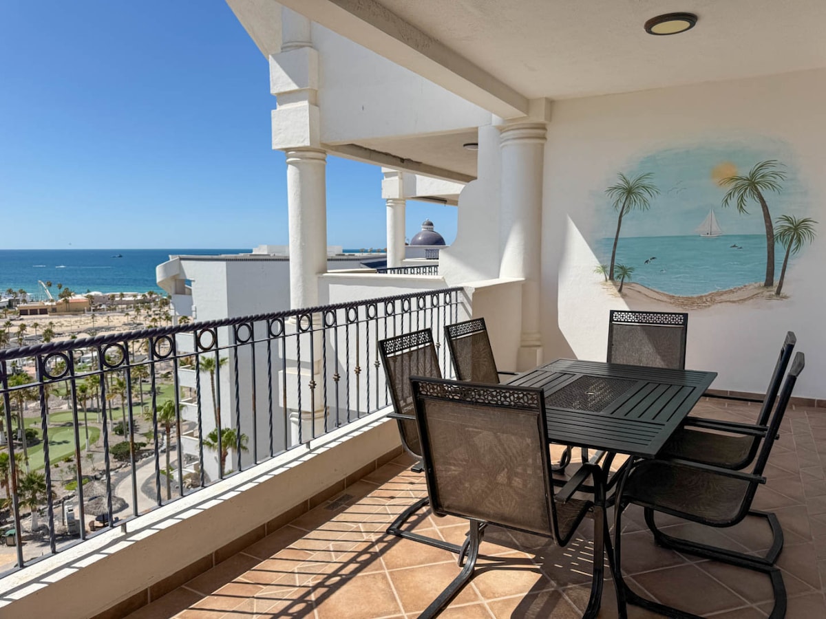 A spacious balcony features a black metal dining table surrounded by four chairs, offering a view of the sea and nearby resort amenities. A decorative mural of a serene beach scene with palm trees enhances the inviting atmosphere.