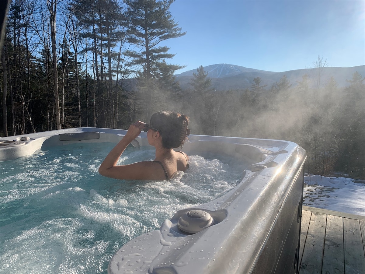 A hydrotherapy hot tub is seen with steam rising from the warm water. A person is relaxing in the tub, surrounded by a natural setting of trees and a distant view of Stratton Mountain under a clear sky.