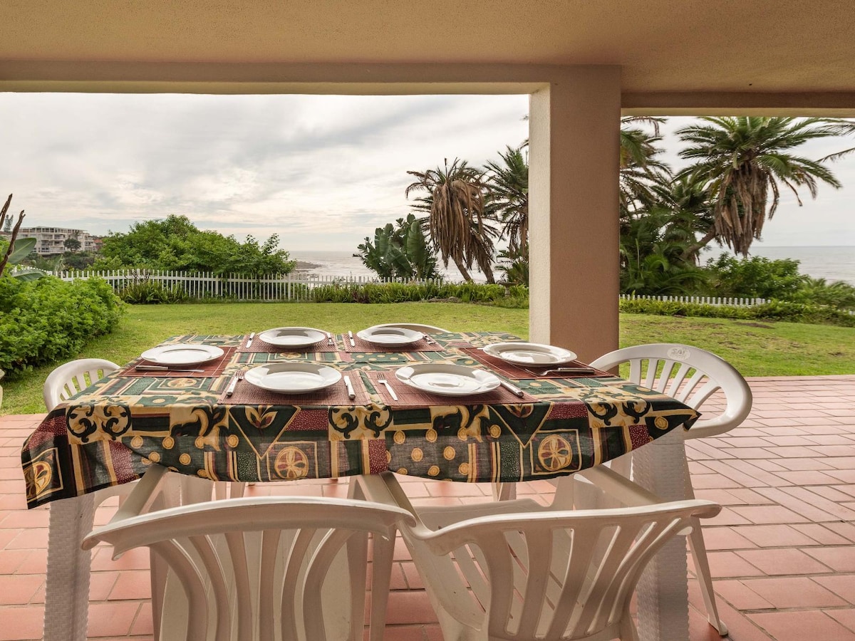 An outdoor dining area is presented with a table set for six, featuring a colorful tablecloth. Palm trees and lush greenery frame the background, with a view of the sea visible beyond the patio. Natural light fills the space, enhancing the relaxed atmosphere.