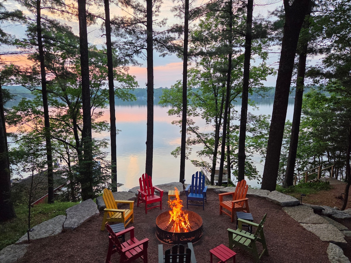 An outdoor fire pit area is surrounded by colorful adirondack chairs, providing a welcoming spot for gatherings. Towering trees frame the serene lake view, while soft hues of sunset reflect on the water's surface, creating a peaceful ambiance.