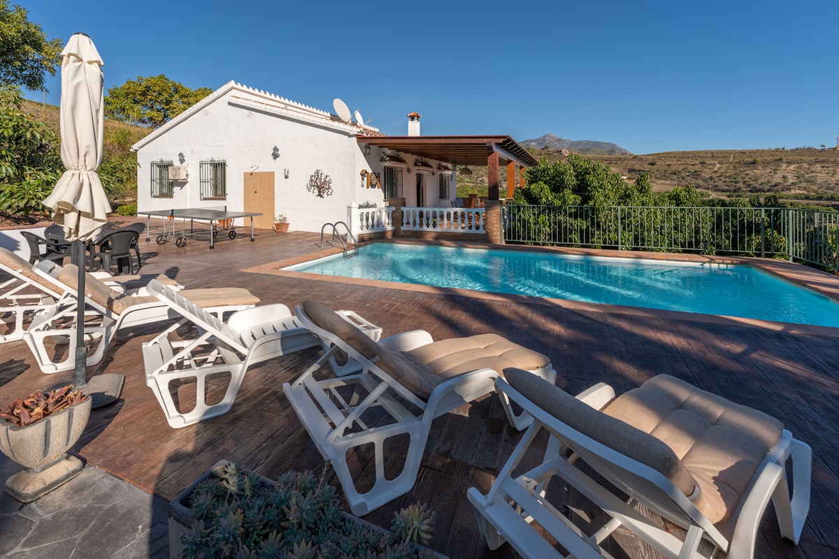An outdoor area features a private pool surrounded by sun loungers and a shaded dining space. Lush greenery is visible in the background, while a whitewashed house and patio offer a relaxed Mediterranean charm.