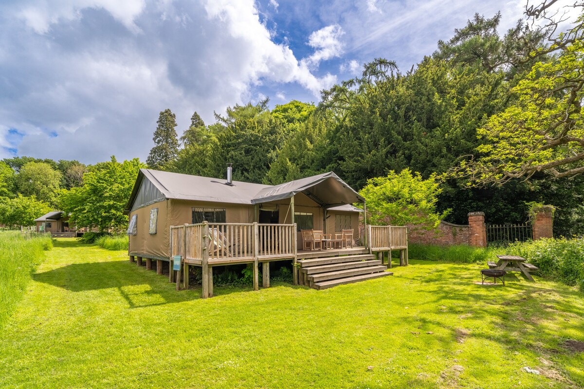 A spacious safari tent is surrounded by lush greenery, set on a wooden deck with outdoor seating. Steps lead from the deck to a grassy area, complemented by trees and a low stone wall in the background under a partly cloudy sky.