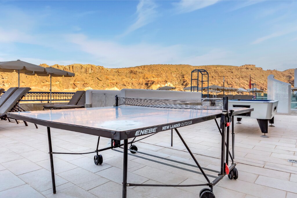 A table tennis table is positioned on a tiled outdoor area, with mountains visible in the background. Several sun loungers, covered by umbrellas, are located nearby, offering a inviting space for relaxation and recreation.