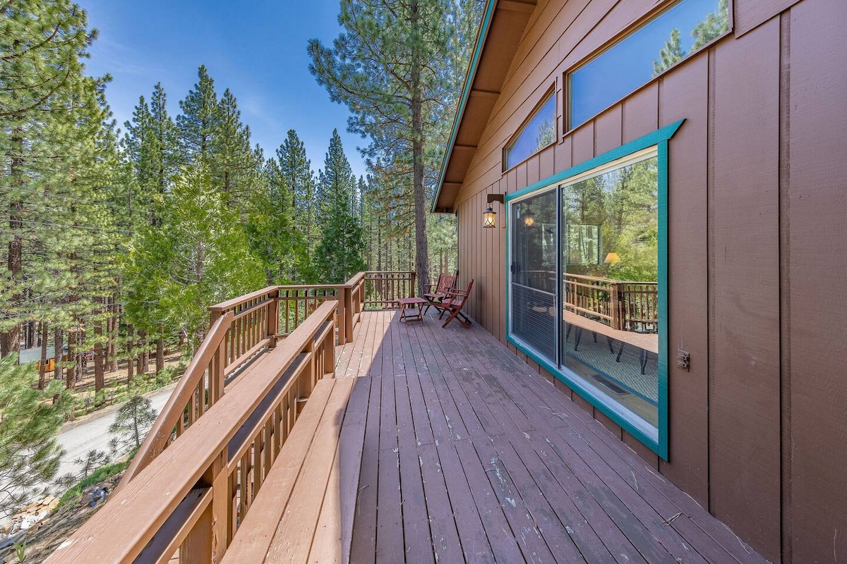 A spacious deck extends from the cabin, surrounded by tall trees and serene woods. Seating areas are present, inviting guests to relax and enjoy the surrounding nature. Large windows reflect the outdoor greenery, allowing natural light to fill the interior space.