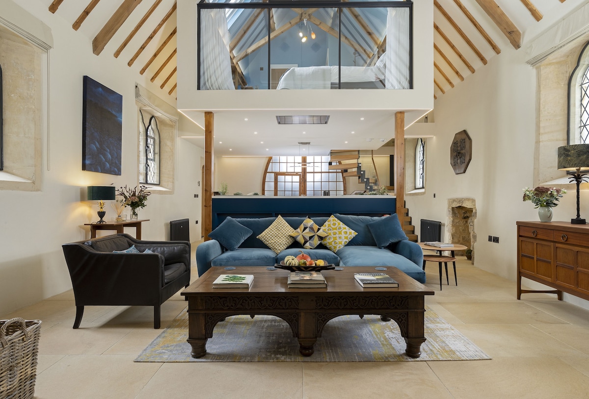 An open-plan living area features a stylish blue sofa adorned with colorful cushions, paired with a dark leather chair and a coffee table. Exposed wooden beams and large windows create a bright atmosphere. A staircase leads to the mezzanine level, enhancing the space's architectural appeal.