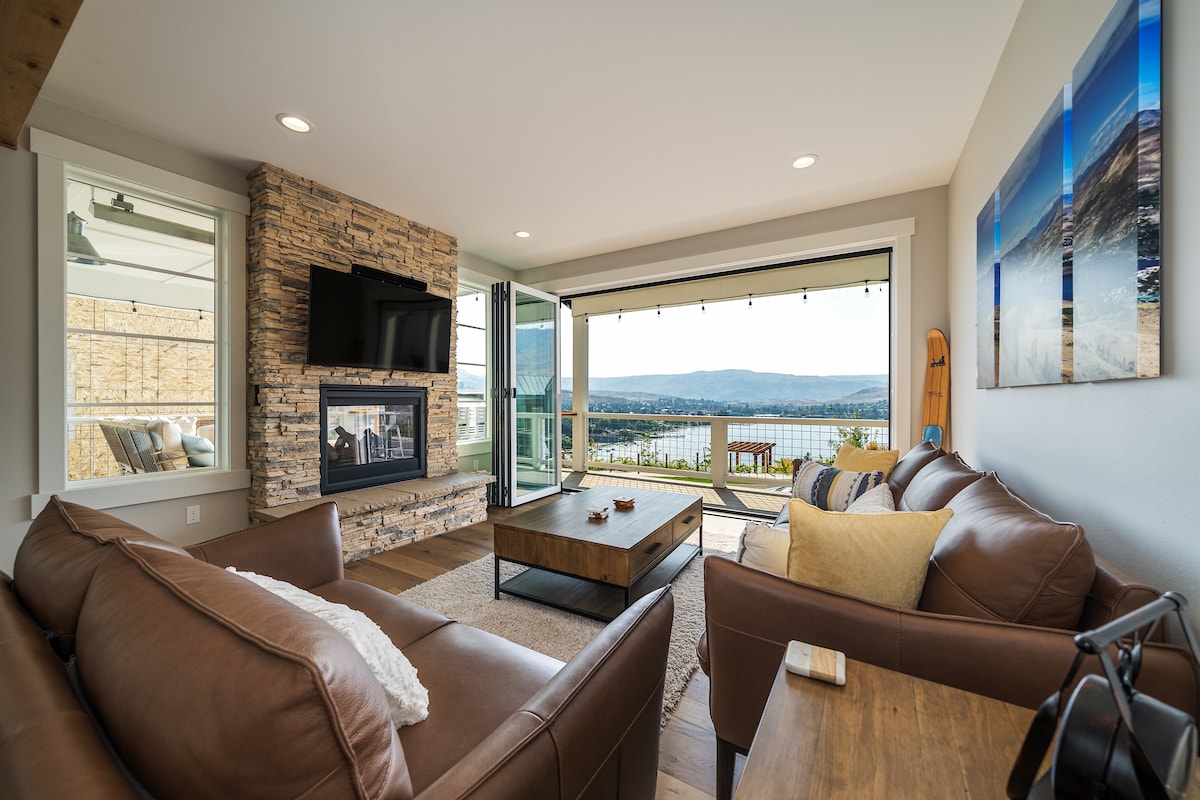 A spacious living area features large glass doors opening to a stunning view of Lake Chelan. A stone fireplace provides a cozy focal point, while comfortable leather sofas and a wooden coffee table invite relaxation. Natural light fills the room, enhancing the welcoming ambiance.