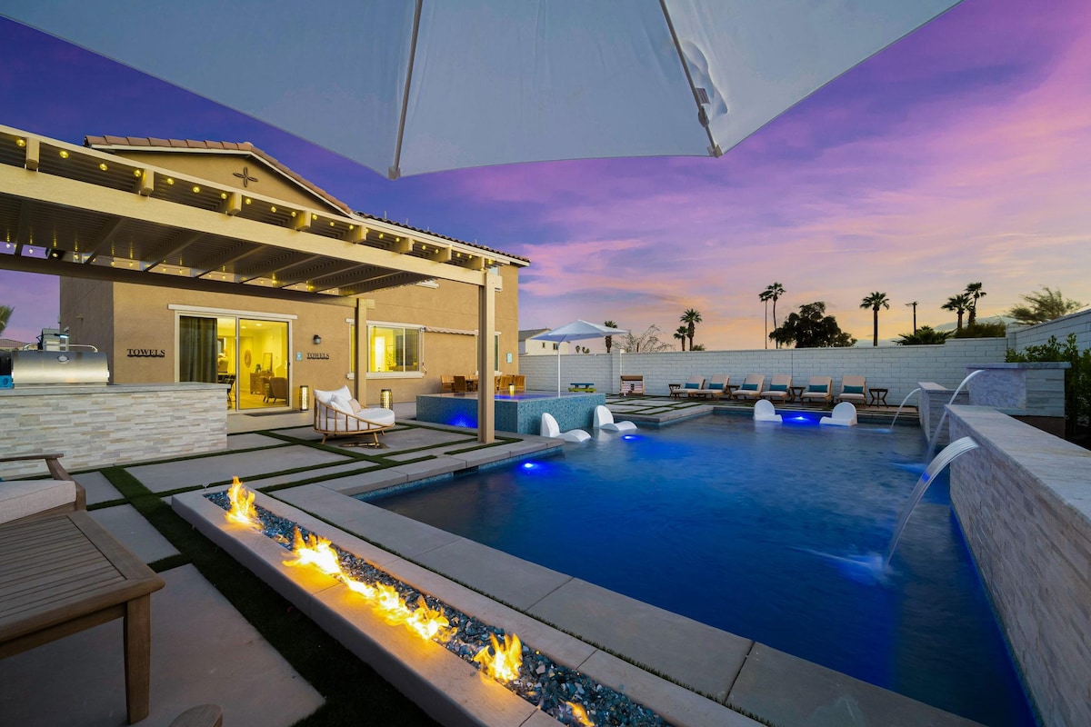 An outdoor relaxation area features a sleek pool with water features and ambient lighting. A spacious patio is accented by lounge chairs and a fire feature, with a backdrop of a professionally designed house illuminated by soft lights. Palm trees frame the scene under a colorful sunset sky.