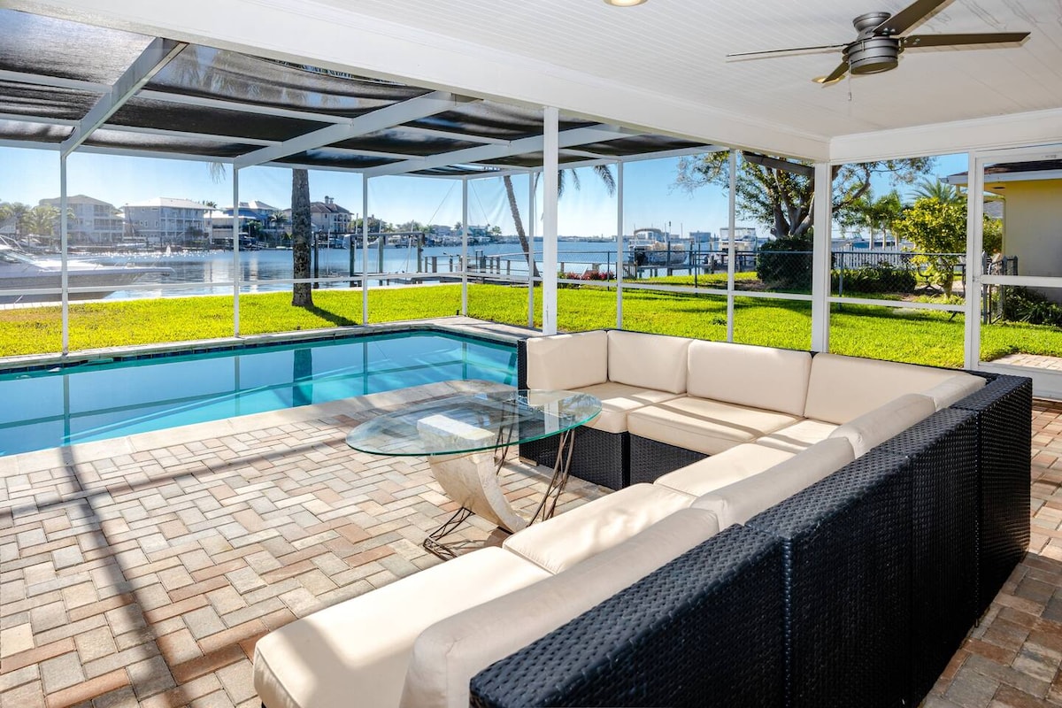 A spacious screened patio overlooks a shimmering pool and a scenic waterfront. A modern sectional sofa encircles a glass coffee table, providing an inviting space for relaxation. Lush green grass and palm trees frame the area, enhancing the outdoor living experience.