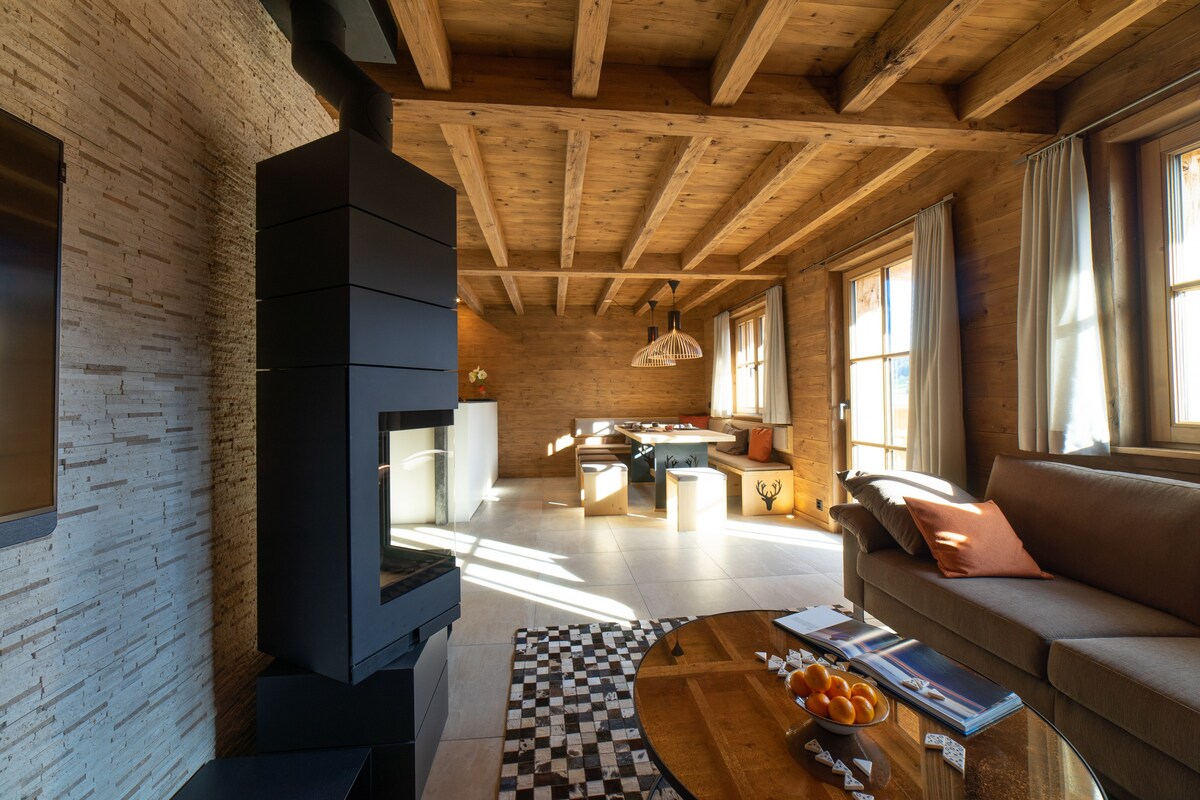 A spacious living area features wooden beams overhead and large windows allowing natural light. A modern fireplace stands on one side, while a comfortable sofa, decorated with accessories, invites relaxation. A circular wooden table is arranged with magazines and oranges, adding a touch of homeliness.