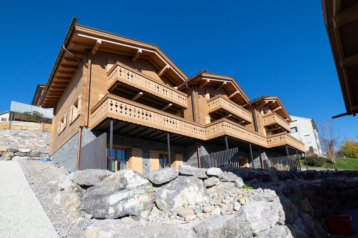 The exterior of the chalet showcases a traditional wooden design, featuring multiple balconies with decorative railings. The structure is nestled against a rocky landscape, complemented by clear blue skies above. A pathway leads to the entrance, enhancing accessibility and integration with the natural surroundings.