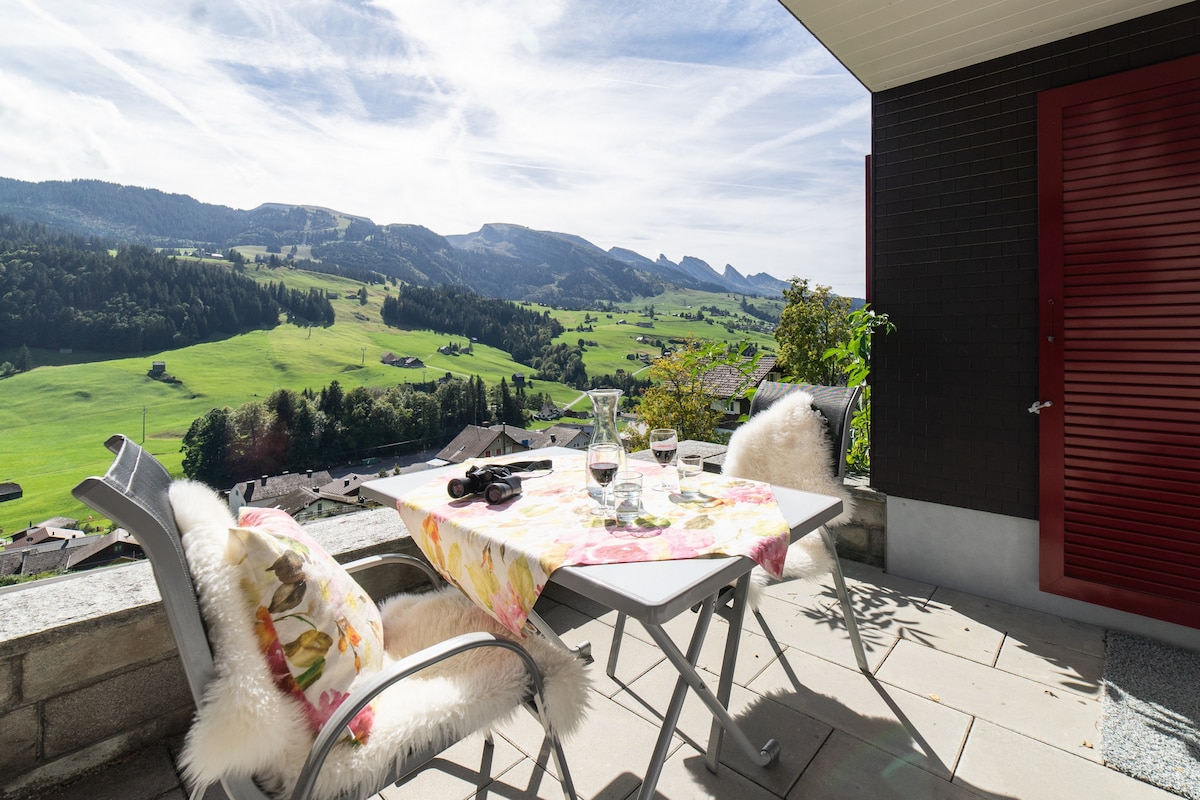 A balcony table is set with drinks and a floral tablecloth, surrounded by chairs. A stunning view of rolling green hills and mountains is visible in the background under a clear sky, creating a serene outdoor space.