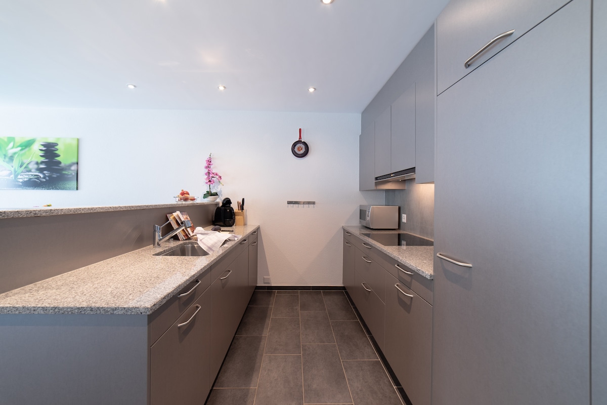 A modern kitchen features sleek gray cabinets and spacious countertops. Equipped with essential appliances, including a microwave and coffee maker, it offers a clean and functional space. Tile flooring adds durability, while soft lighting enhances the overall atmosphere.