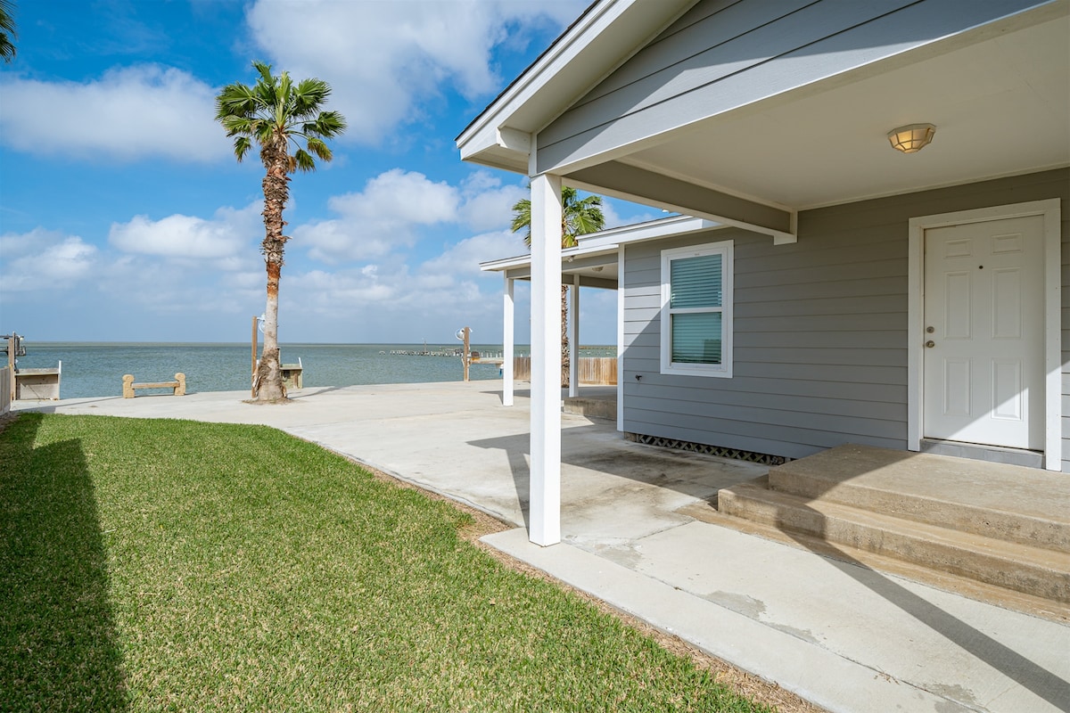 The exterior view showcases a coastal home with grey siding, a light-colored front door, and a stoop leading to the entrance. A well-manicured lawn and palm trees frame the property, complemented by a clear blue sky and serene water visible in the background.