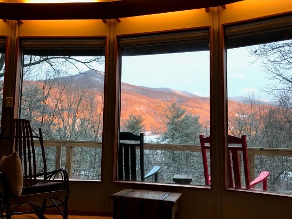A curved wall of windows frames a serene view of the landscape outside. The hills are highlighted by warm light, and two colorful chairs in red and blue are positioned on the deck, inviting relaxation while taking in the natural surroundings.