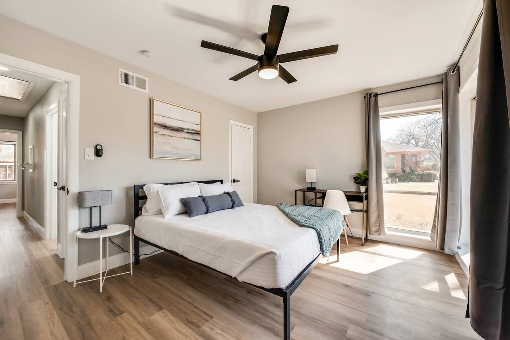 Një dhomë gjumi moderne me një krevat dopio "queen" me shtroja të bardha dhe një batanije. Një ventilator tavani me një ndriçues të integruar është pozicionuar mbi krevat. Një tavolinë pranë krevatit me një abazhur është ulur në të majtë.
