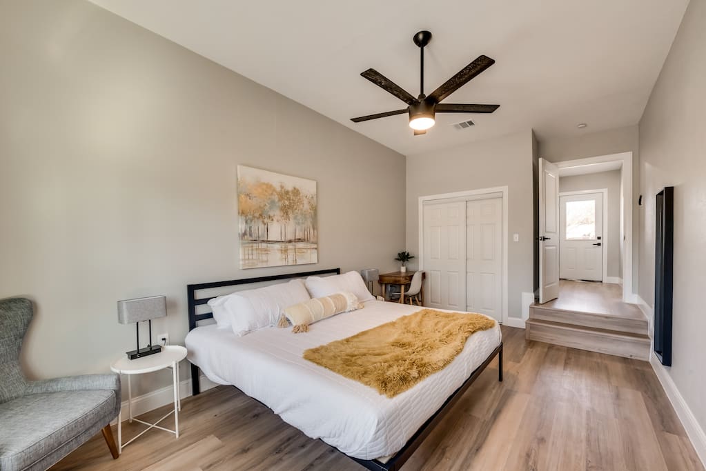 Një dhomë gjumi e rehatshme me një krevat qendror me shtroja të bardha dhe një batanije me ngjyrë mustarde. Një ventilator tavani me tehe të zeza është montuar mbi krevat. Një komodinë me një abazhur qëndron në të majtë, ndërsa një pikturë shfaqet mbi kokë.

