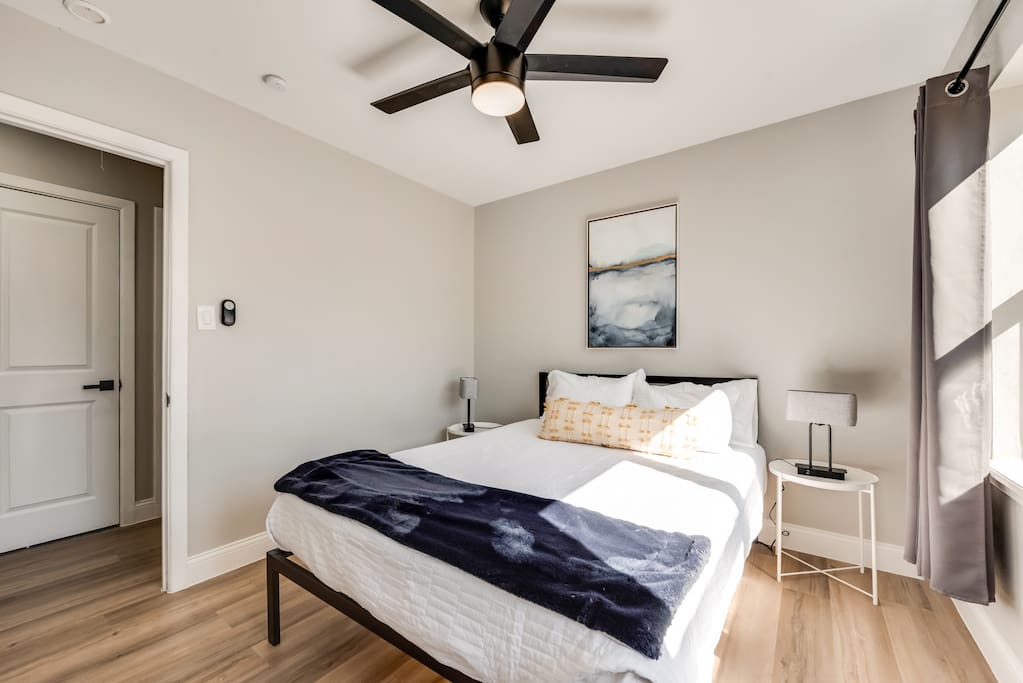 Një dhomë gjumi moderne me shtroja të bardha, një hedhje blu e errët, vepra arti abstrakte sipër, një ventilator tavani dhe një tavolinë pranë krevatit.