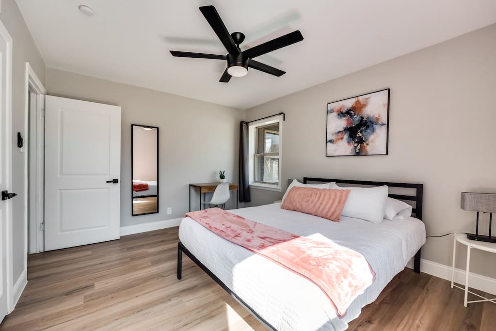 Një dhomë gjumi elegante me një krevat dopio "queen" me shtroja të bardha dhe një batanije rozë. Krevati është në qendër me një jastëk me theks të modeluar që shton karakter. Mbi të, një ventilator i zi tavani me një ndriçues e ankoron dizajnin. 