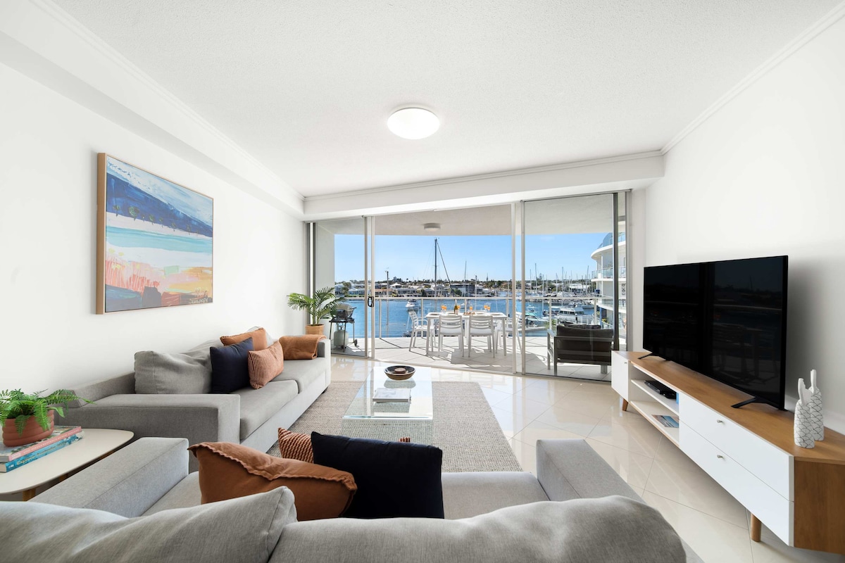 The open-plan living area showcases large glass doors that provide stunning views of the marina. A comfortable seating arrangement with neutral-toned sofas faces a modern TV unit, while a coffee table sits in the middle. The adjacent outdoor balcony features a dining setting for alfresco meals.