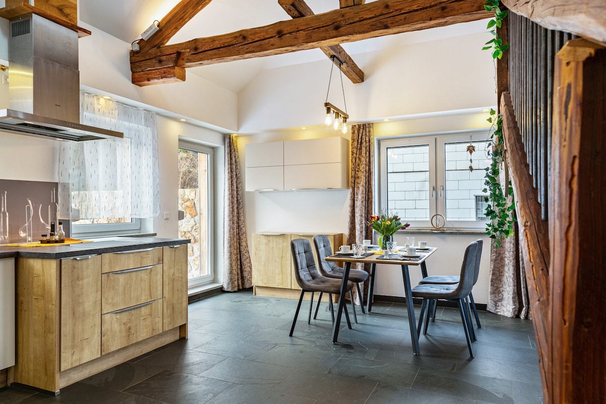 A modern kitchen and dining area features wooden beams overhead, providing a rustic charm. The well-equipped kitchen includes sleek cabinetry and modern appliances. A dining table is arranged for four, complemented by elegant chairs, with natural light flowing through large windows.