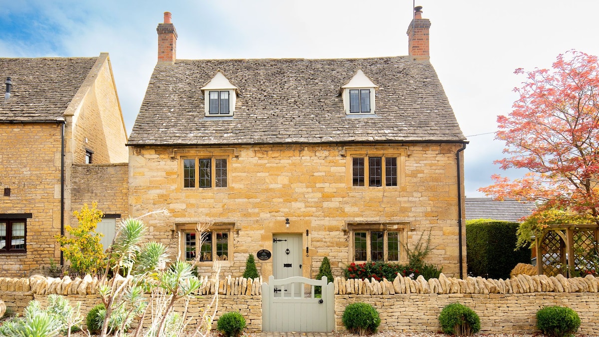 The charming Cotswold stone cottage is showcased, with a well-maintained façade featuring large windows and a welcoming door. A small front garden is bordered by a low stone wall, complemented by a tidy pathway and a pergola draped in greenery on one side.