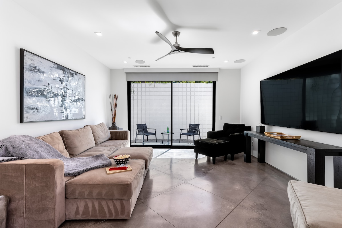 A modern living area features a comfortable sectional sofa with a throw blanket and a side table. Floor-to-ceiling glass doors open to an outdoor space. A flatscreen TV is mounted on the wall, while a sleek console table and a dark accent chair complete the setup.