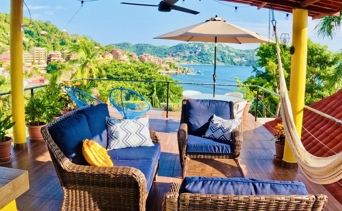 A rooftop patio features outdoor seating with two chairs and a loveseat, set against scenic views of the ocean and surrounding hills. A large umbrella provides shade over the seating area, while a hammock is hung nearby, inviting relaxation amidst lush greenery.