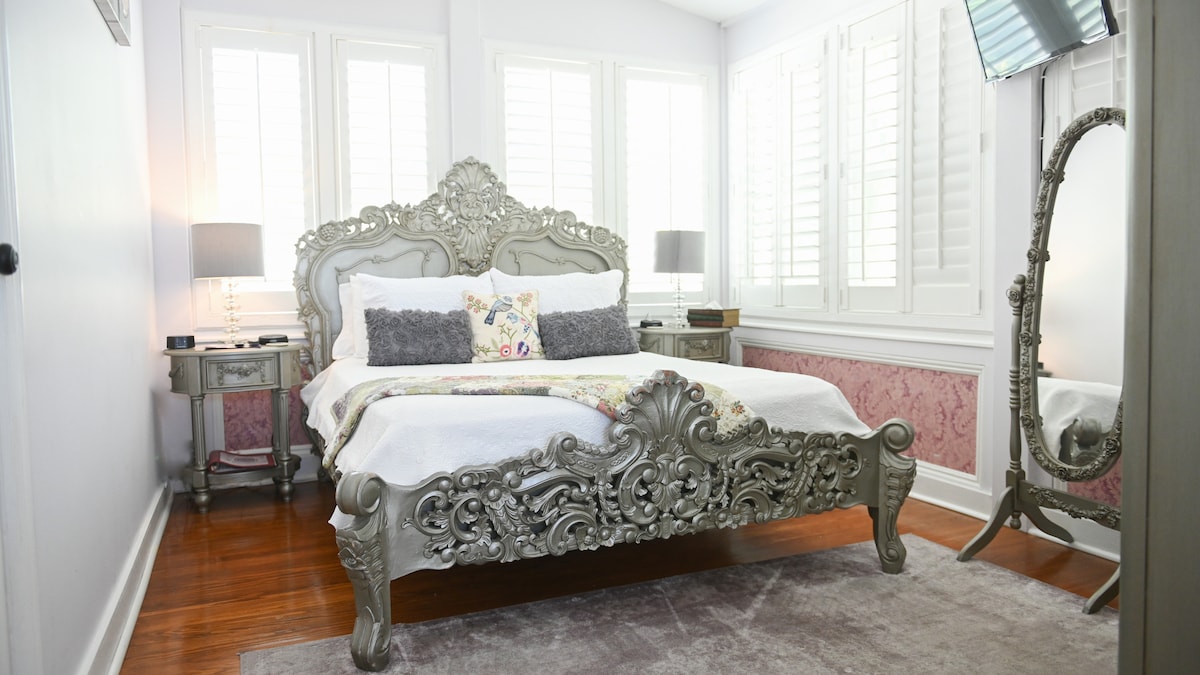 A queen-sized bed with intricate, ornate detailing is positioned against a soft grey wall. Natural light filters through Bahamian shutters, illuminating the room's elegant decor. A vintage-style mirror stands nearby, and two bedside tables with lamps complement the chic Parisian ambiance.