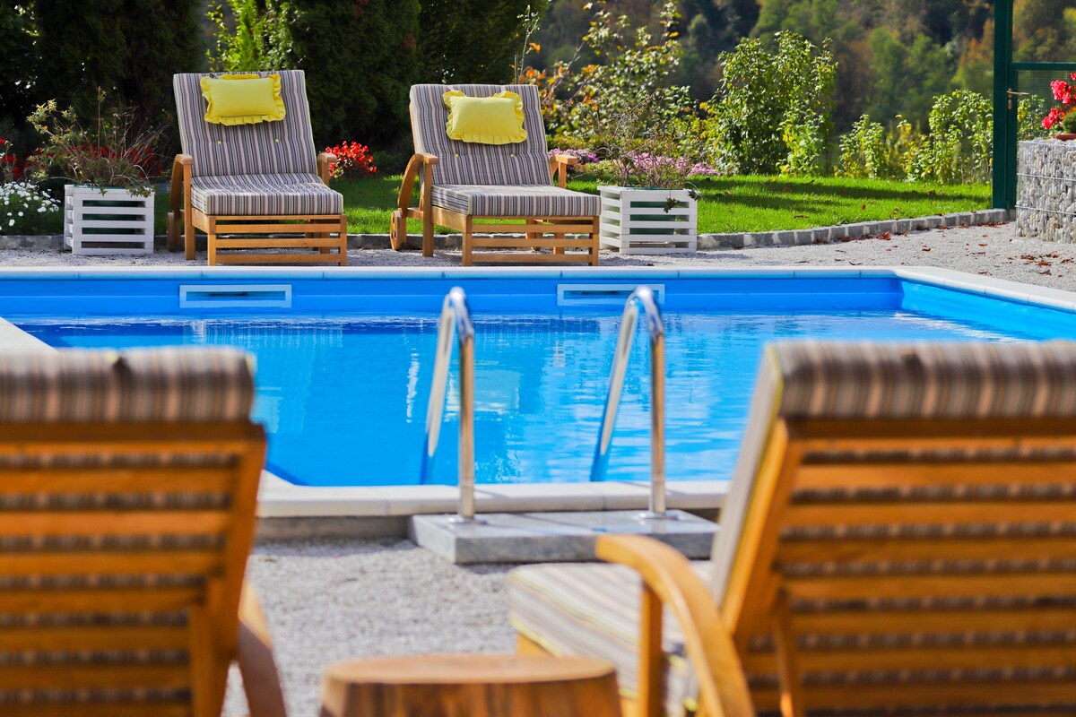 A refreshing private pool is highlighted by the clear blue water and surrounded by a well-maintained garden. Two wooden loungers with striped cushions are positioned nearby, inviting relaxation. Colorful flowers in planters enhance the natural scenery, creating a serene outdoor space.